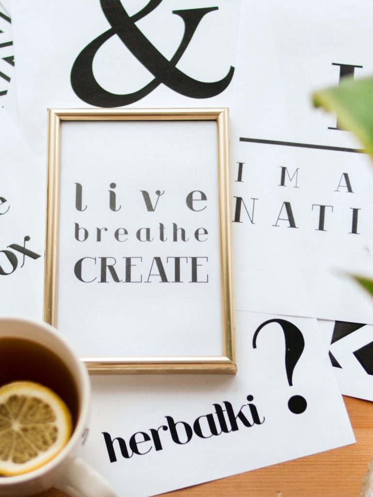 Motivational quote in a golden frame reading 'Live Breathe Create' placed on a desk with tea and typography sheets, symbolizing creativity and wellness.